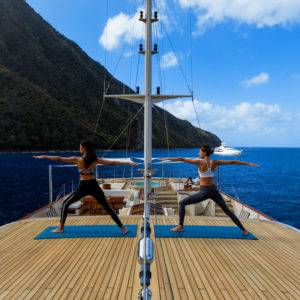Two women in athletic wear doing yoga on the top deck of NERO Yacht with grassy mountain and boat off in the distance.