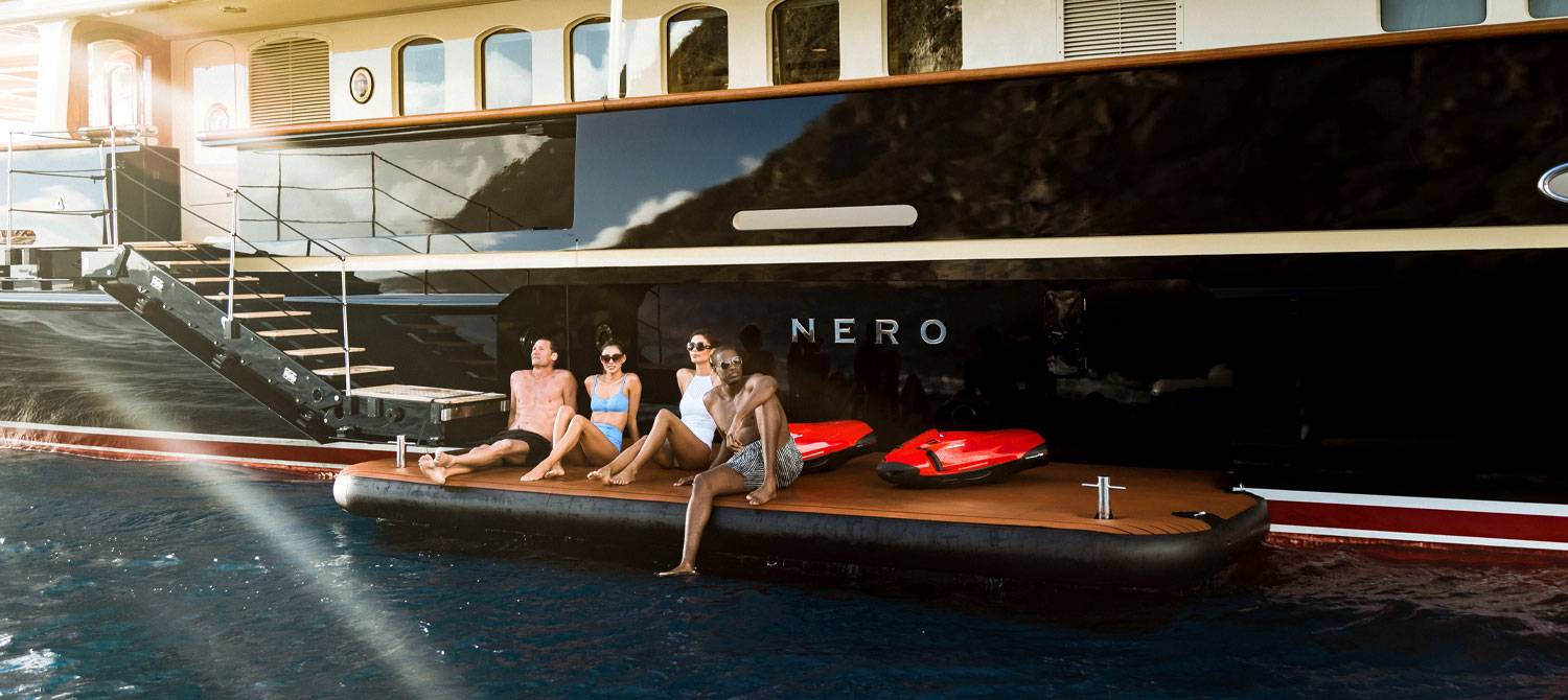 Two men and two women in bathing suits sitting on the swim platform of NERO yacht with red SeaBobs