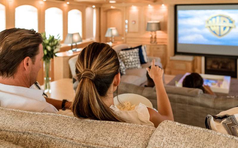 Back of a man and woman's heads, sitting on a couch. Woman is pointing at a TV screen with Warner Brothers logo on it.