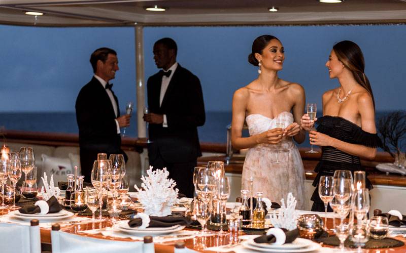 Two men in tuxedos and two women in strapless formal gowns standing behind a long set table on NERO Yacht.